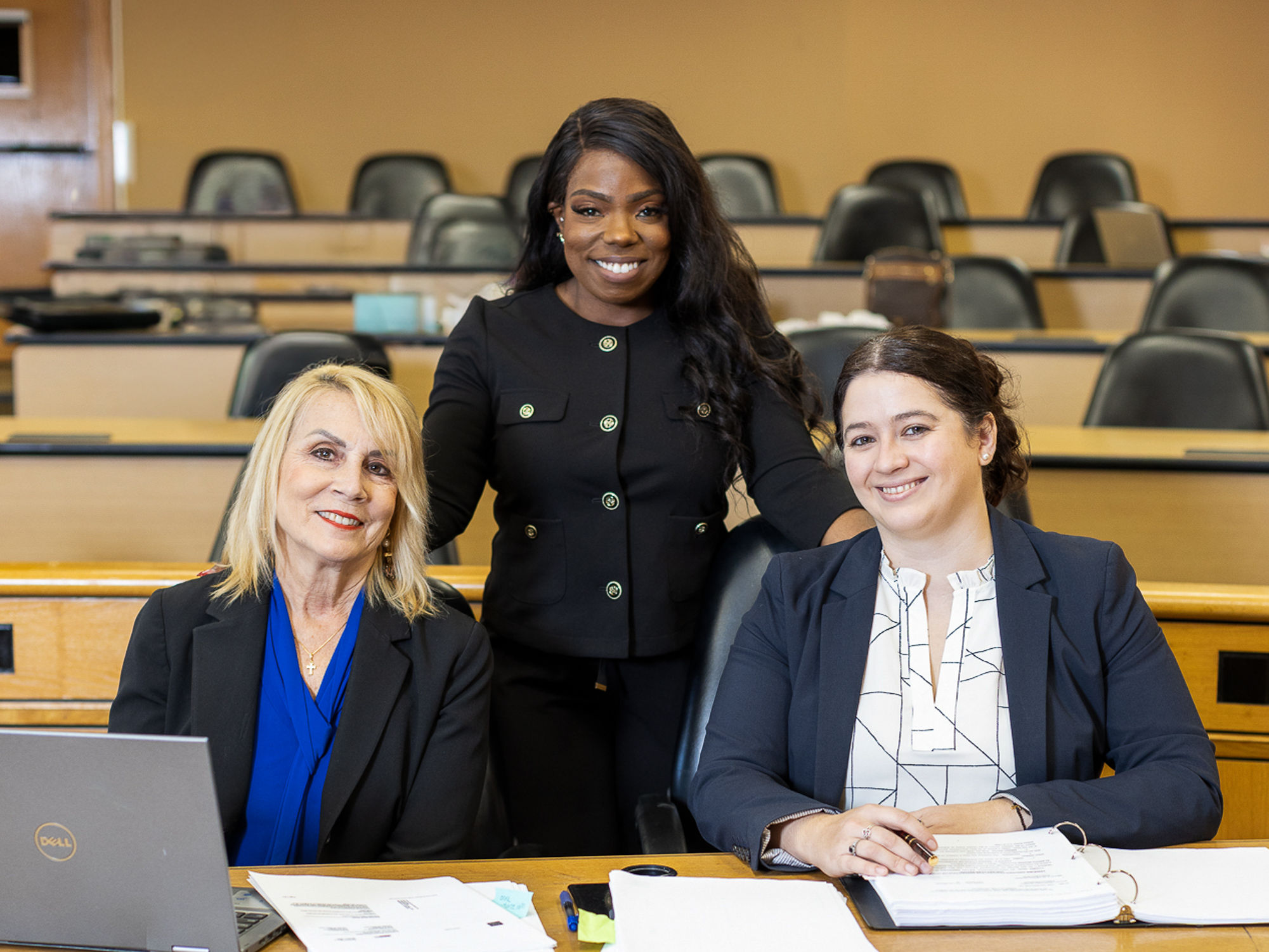 Three lawyers posing in a court room, one standing behind the other two seated