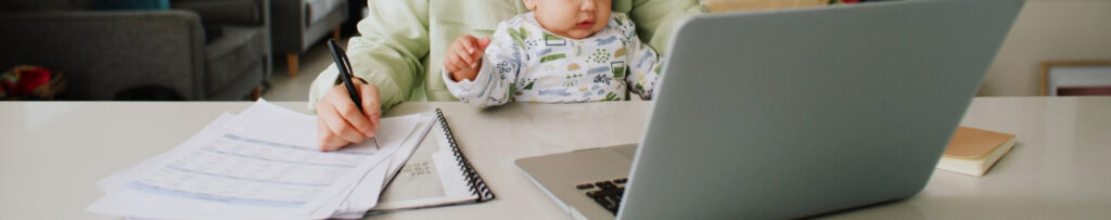 woman holding newborn on lap at desk looking at lap top and writing
