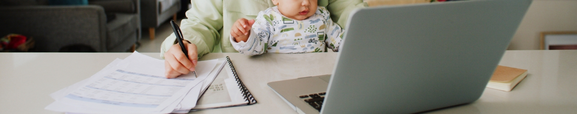 woman holding newborn on lap at desk looking at lap top and writing